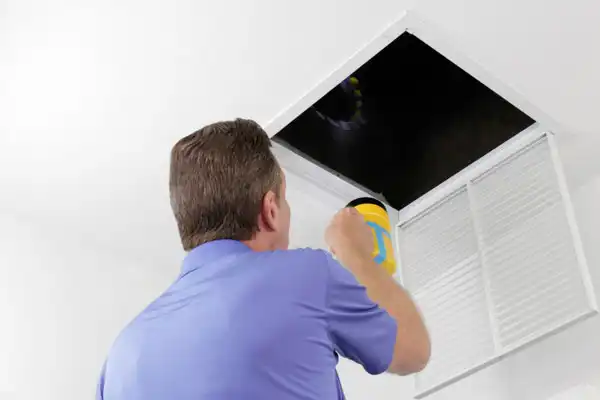 A person in a blue shirt holds a flashlight, looking into an open ceiling vent in a white room.