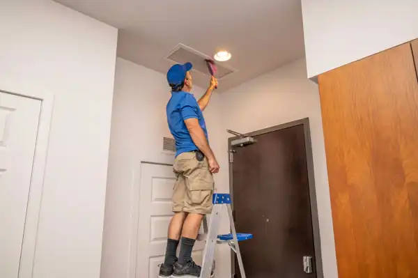 Person on a ladder cleaning a ceiling air vent with a brush, wearing a blue shirt, cap, and beige shorts, in a room with white walls and a wooden door.