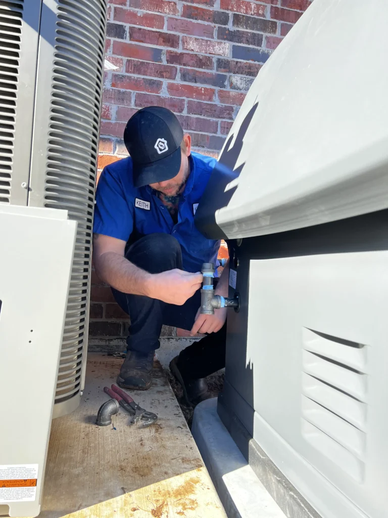 Technician in a blue uniform and cap works on a grey generator unit next to another large machine, with brick wall in background.