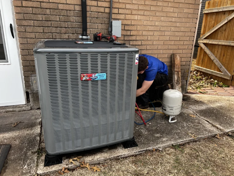A technician in a blue shirt works on a large outdoor HVAC unit next to a brick wall, with tools and a propane tank nearby.