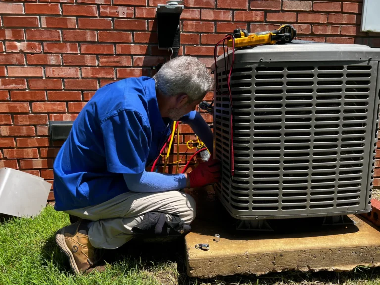 Technician in a blue shirt and red gloves repairs an outdoor air conditioning unit against a brick wall.