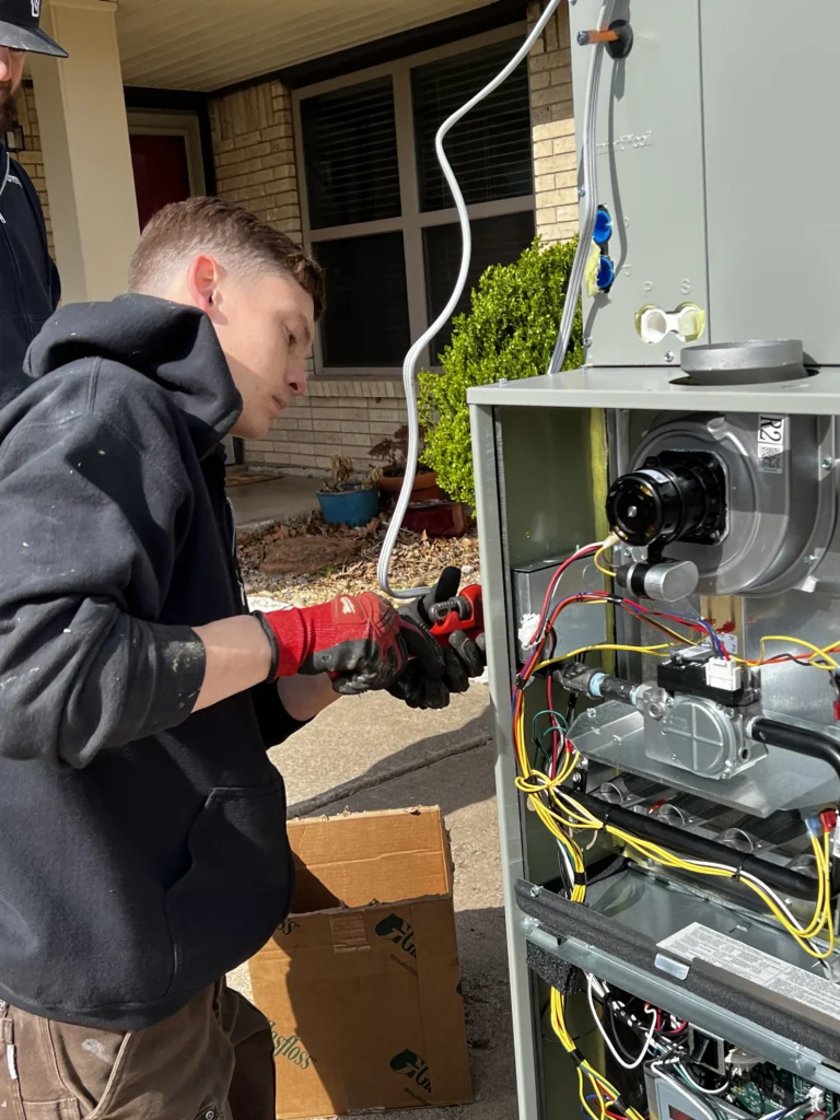A person in a black hoodie and red gloves works on a furnace with visible wires and components outside a house.
