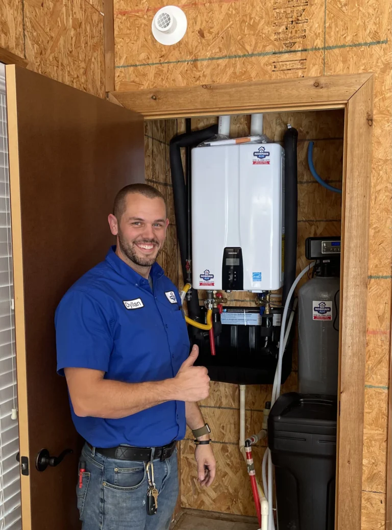 A man in a blue shirt gives a thumbs up next to a white wall-mounted water heater in a wooden enclosure.
