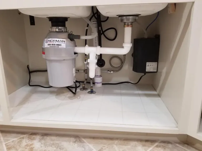 View under a kitchen sink showing plumbing pipes, a garbage disposal unit labeled "NDRMAN," and various connections. The area is clean and neatly organized.