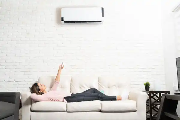 A woman reclines on a white couch, pointing a remote at a wall-mounted air conditioner in a room with a white brick wall.