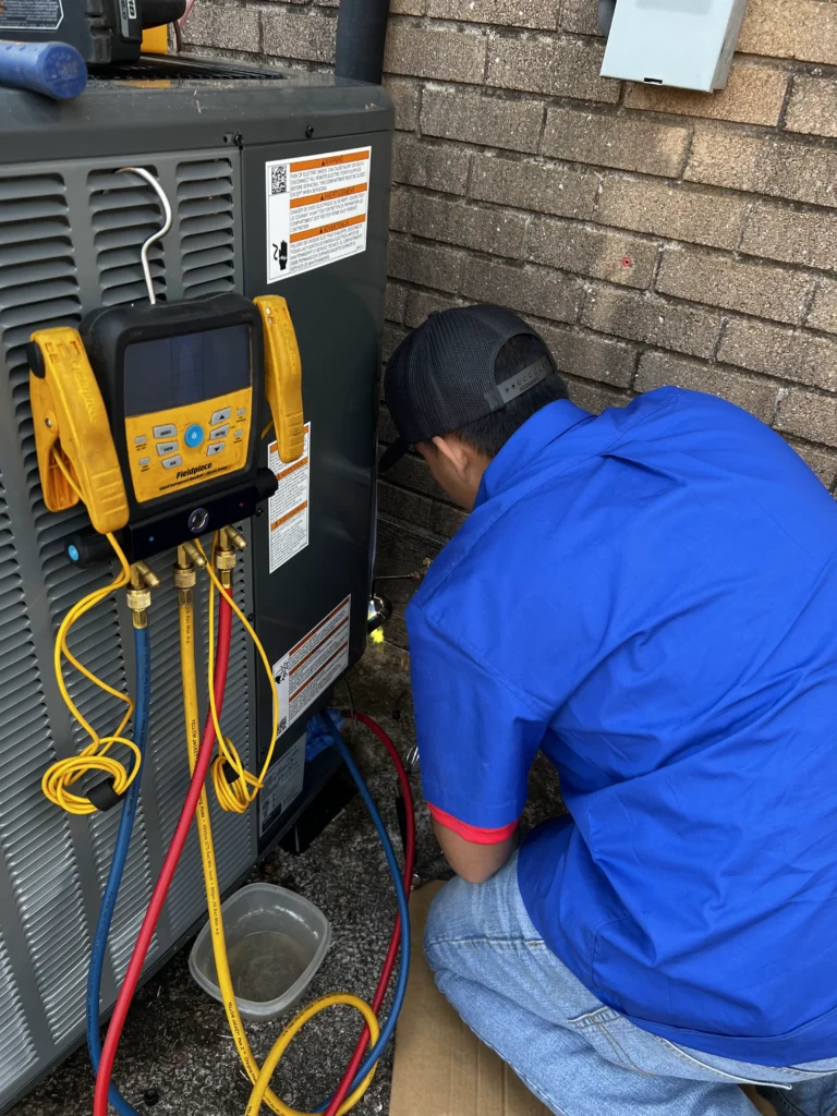 Technician in blue shirt and cap works on an outdoor HVAC unit, with yellow, blue, and red hoses connected to gauges by a brick wall.