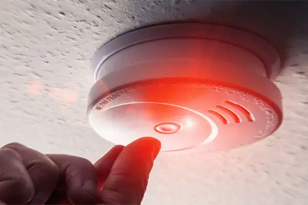 A person tests the detector installation by pressing the button on a ceiling-mounted smoke detector, its red light glowing ominously.