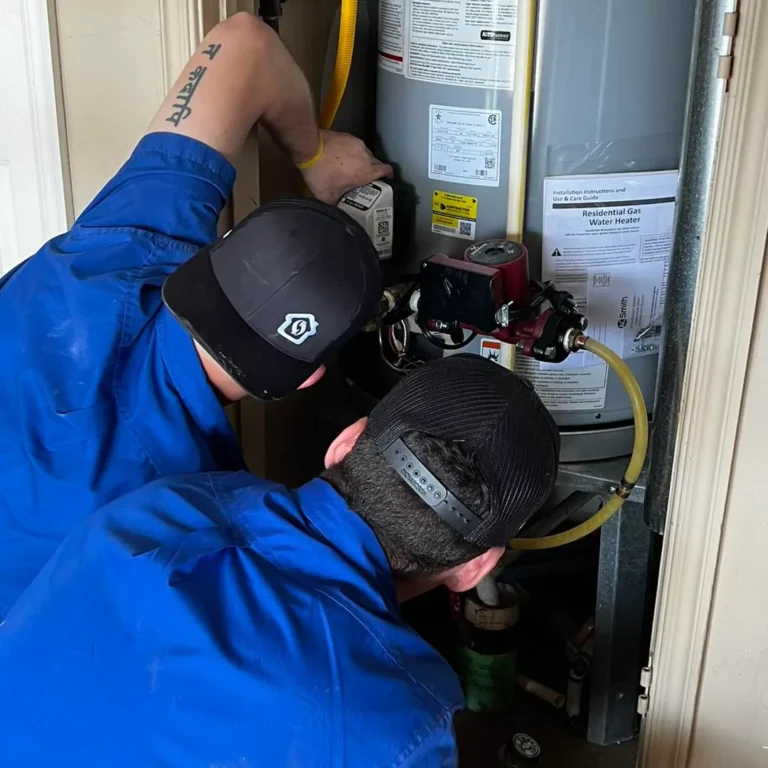 Two workers in blue uniforms and caps are inspecting a water heater unit in a confined space, focusing on its components and connections.