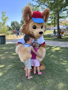 A large furry costume character, embodying the spirit of joy and connection that defines us, embraces two young children outdoors on a grassy area near tents and trees.