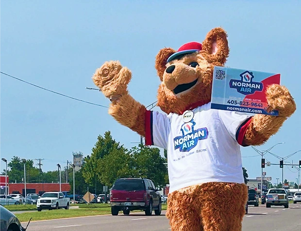 Person in a bear mascot costume wearing a "Norman Air" T-shirt and hat holds a company sign while waving at cars on a street.