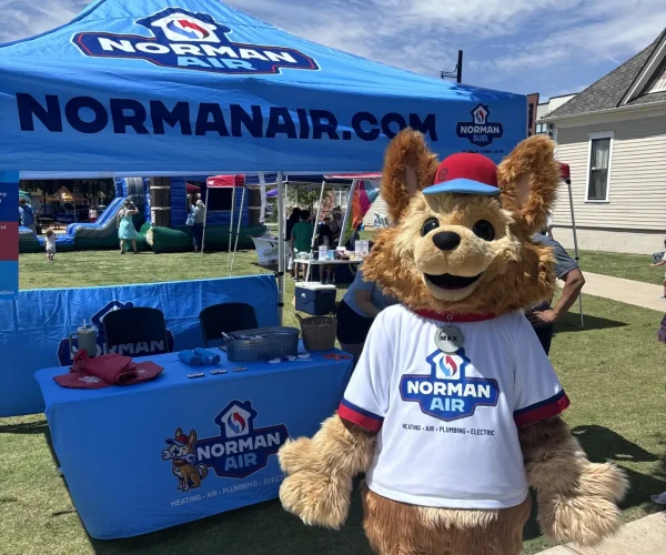 Max tent A mascot dressed as a dog stands proudly in front of a Norman Air tent at an outdoor event, inviting attendees to learn more about us. The tent is bustling with promotional materials and a welcoming table, while the sky above is clear with a sprinkling of clouds.